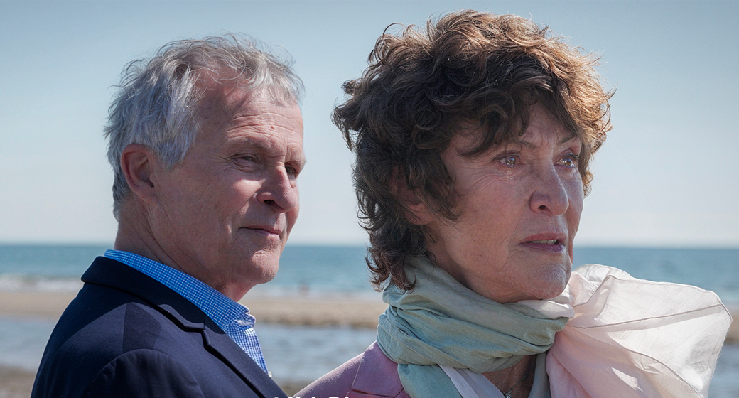 Photo d'une plage avec en premier plan un homme âgé de profil et devant lui une femme agée en train de pleurer. Devant eux, au centre le logo textuel de la "Un Si Grand Soleil"