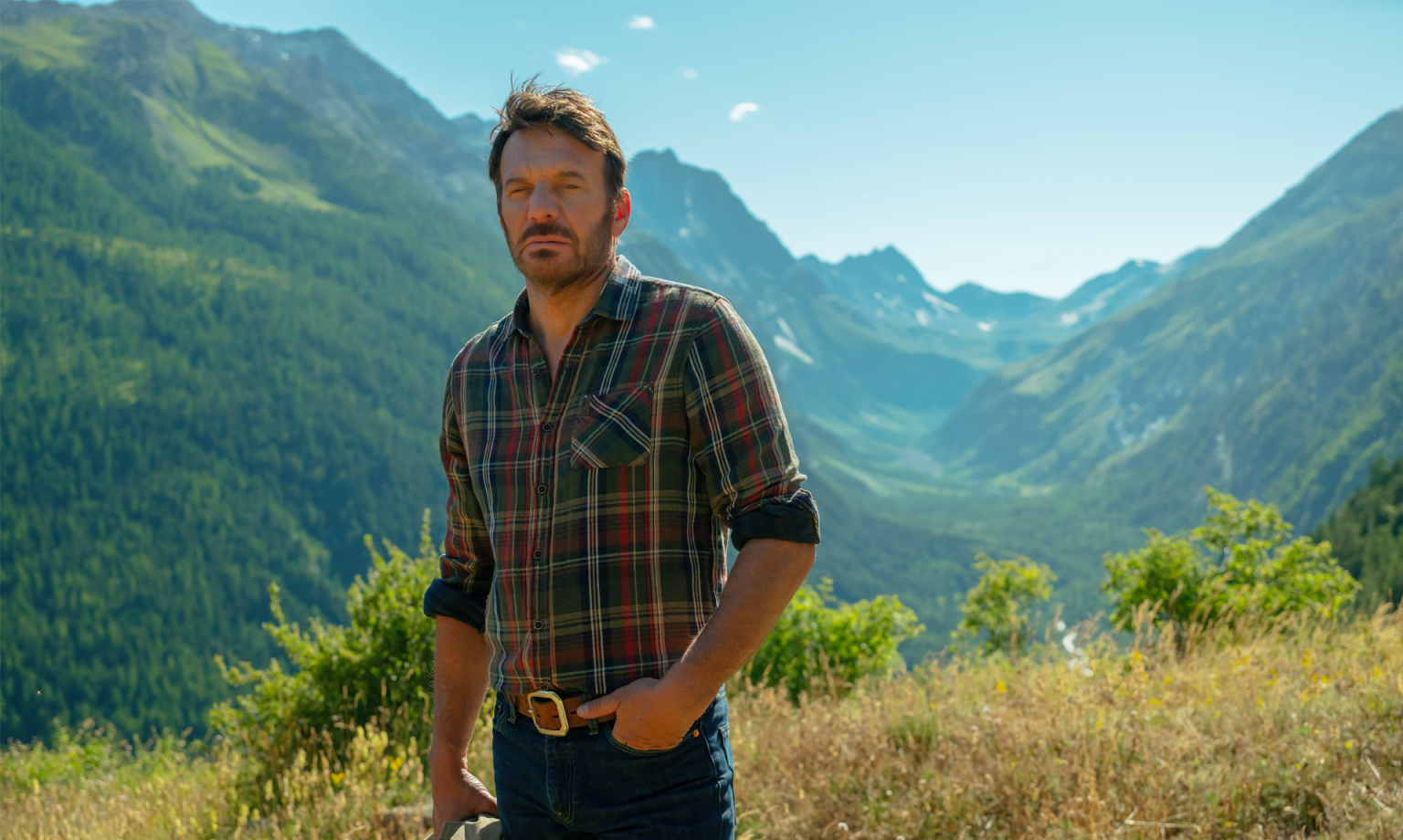 Alex Hugo (Samuel Lebihan) debout de face, derrière lui une belle vue verdoyante de montagne