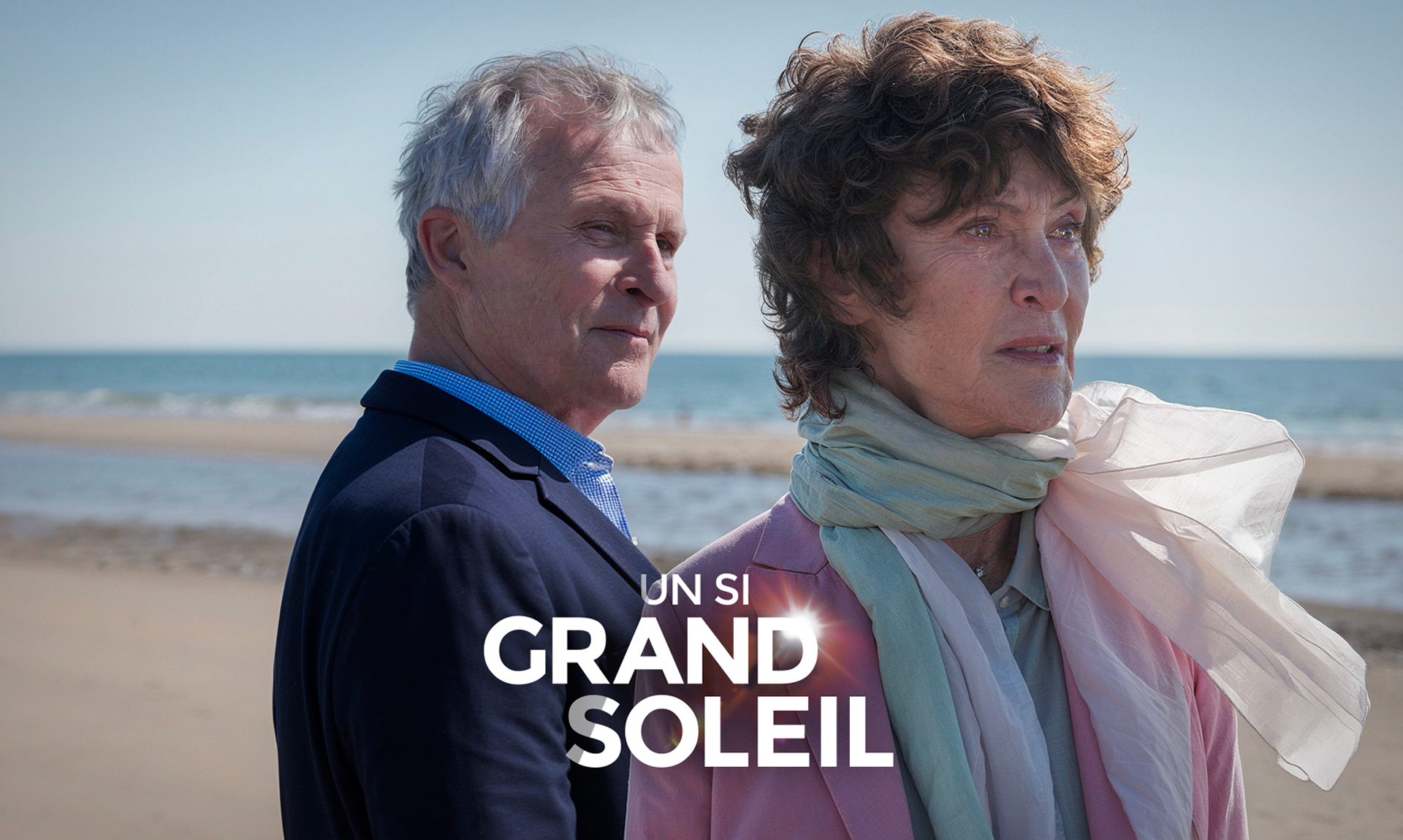 Photo d'une plage avec en premier plan un homme âgé de profil et devant lui une femme agée en train de pleurer. Devant eux, au centre le logo textuel de la "Un Si Grand Soleil"