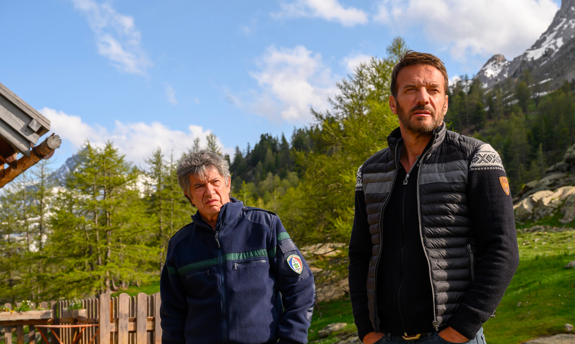 Samuel Le Bihan, dans le rôle d'Alex Hugo, se tient aux côtés d'un garde forestier dans un décor alpin. Les deux hommes observent attentivement les environs, entourés de prairies, de conifères et de montagnes partiellement enneigées sous un ciel lumineux. Cette scène reflète l'ambiance naturelle et l'univers des enquêtes de la série Alex Hugo.