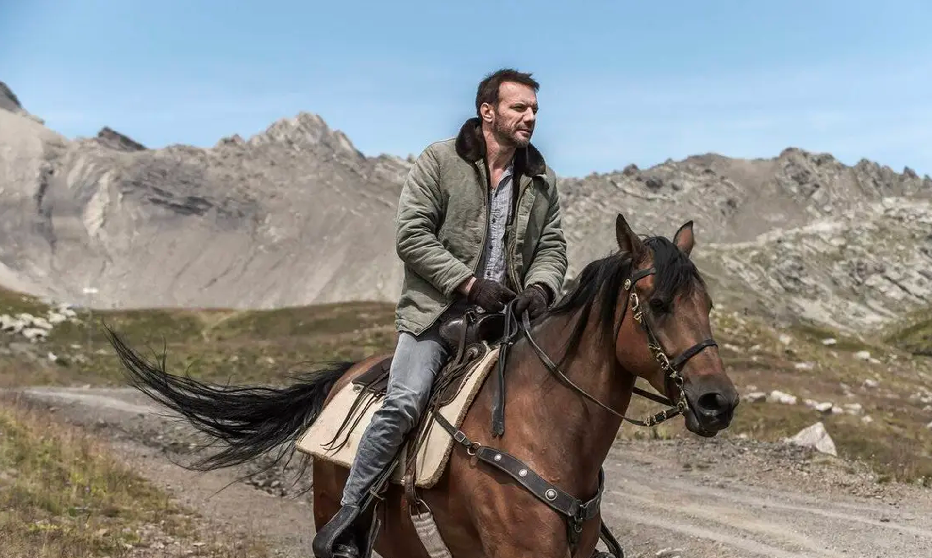 Samuel Le Bihan, interprète du personnage d'Alex Hugo, chevauche un cheval brun sur un sentier de montagne. Il porte une veste verte doublée, des gants et un jean, tandis qu'un paysage de sommets rocheux s'étend à l'arrière-plan sous un ciel bleu. Cette scène illustre l'atmosphère sauvage et contemplative de la série Alex Hugo.